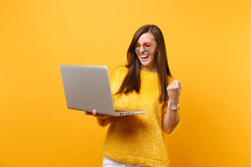 Laughing young woman in sweater, heart eyeglasses doing winner gesture working on laptop pc computer isolated on bright yellow background. People sincere emotions, lifestyle concept. Advertising area.