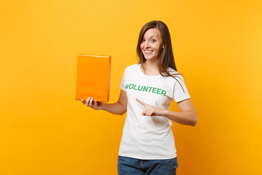 Portrait of smiling woman in white t-shirt with written inscription green title volunteer hold orange cardboard box isolated on yellow background. Voluntary free assistance help, charity grace concept