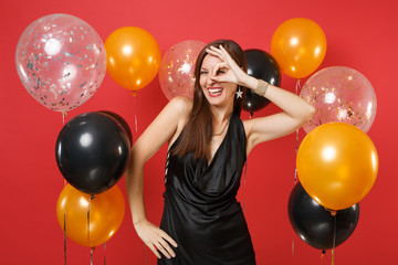 Laughing young woman in little black dress celebrating, showing OK sign near eyes on bright red background air balloons. St. Valentine's Day, Happy New Year, birthday mockup holiday party concept.