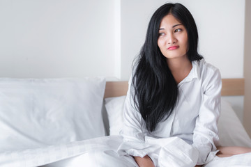 Beautiful young pretty Asian woman wake up and make happy smile with white shirt at the white bed in the morning 