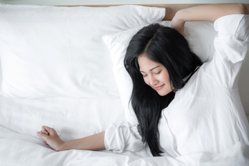 Beautiful young pretty Asian woman wake up and make happy smile with white shirt at the white bed in the morning 