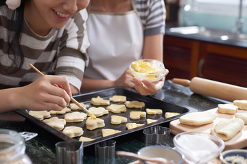 Close-up view of anonymous women applying butter on raw cut out cookies  lying on baking tray in home kitchen