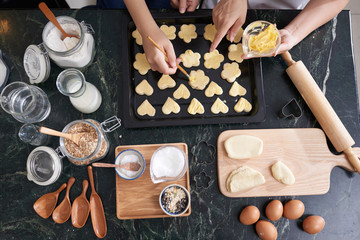 Directly above of female hands spreading butter on uncooked cookies lying on baking tray over kitchen table with ingredients and cookware