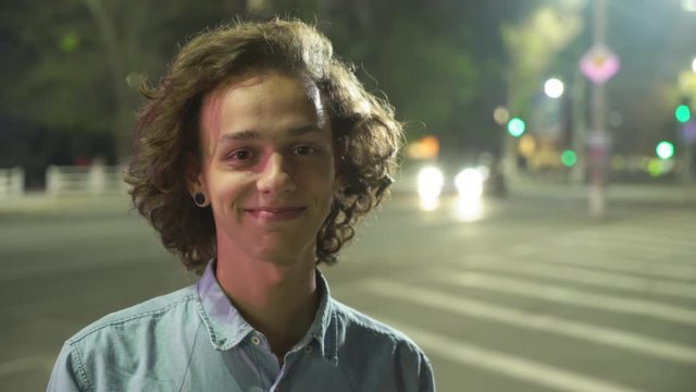  Portrait of a glamour man with long wavy hair standing at zebra crossing with traffic and smiling openheartedly in a street at night in autumn  