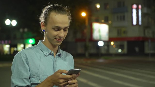  Profile of a cheery young man with a bun haircut surfing the net on his smartphone, smiling and seeking his sweetheart rin a street at night in autumn