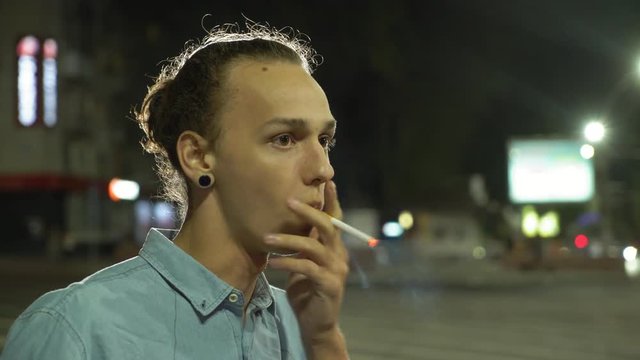  Portrait of a glamour young man with a bun hairstyle and earrings is lighting a cigarette ann smoking in a city street at night in autumn