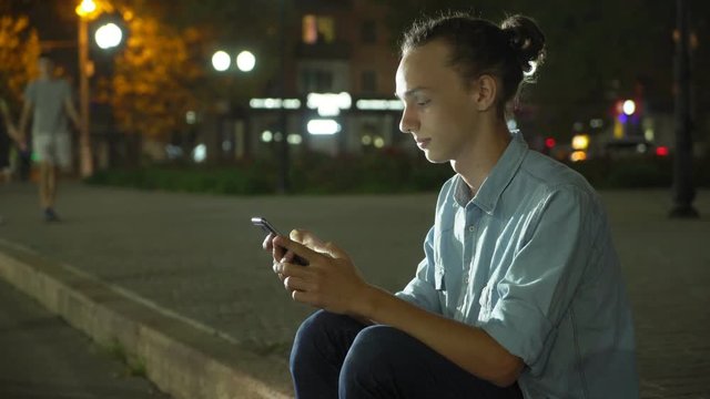  Profile of an inspired young man with a bun haircut browsing the net on his smartphone and seeking info in a city street deep at night in autumn
