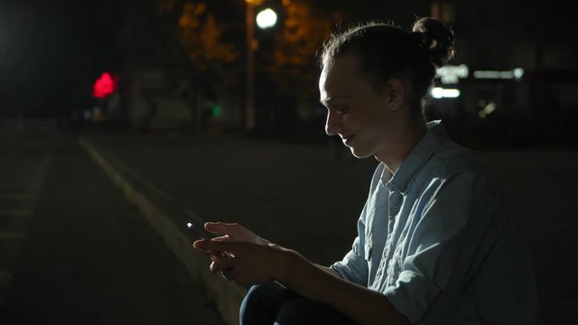  Profile of a smiling young man with a bun hairstyle surfing the net cheery on his smartphone and seeking photos in a green park deep at night in autumn