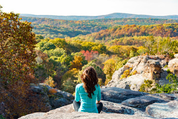 Woman in yoga pose thinking and meditating on cliff overlooking  landscape during the day sunrise or sunset changing fall leaves with orange yellow green brown and red leaves