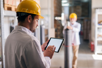 Picture of young focused male manager standing in factory and holding tablet. Dressed in white coat...