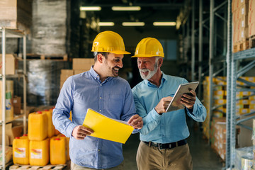 Two successful smiling business man walking through big warehouse with helmets on their heads....