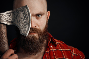 bearded man with mustache is holding a battle axe cover his eye and looking on camera on black...