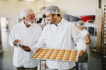Picture of two employees in sterile clothes in food factory smiling and talking. Younger man is holding tray full of fresh cookies while the older is holding tablet and checking production line.