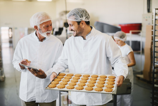 Picture of two employees in sterile clothes in food factory smiling and talking. Younger man is holding tray full of fresh cookies while the older is holding tablet and checking production line.