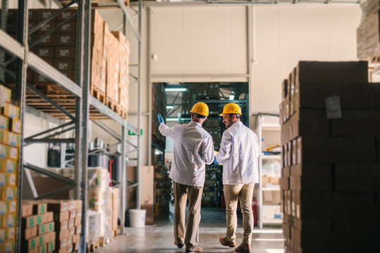 Father and son standing in their warehouse with helmets on their heads and looking at package prepared for transport. Looking proud and satisfied.