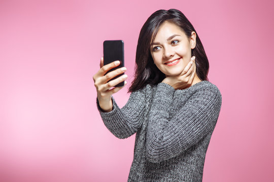 Portrait of attractive young woman taking selfie on smartphone on pink background.