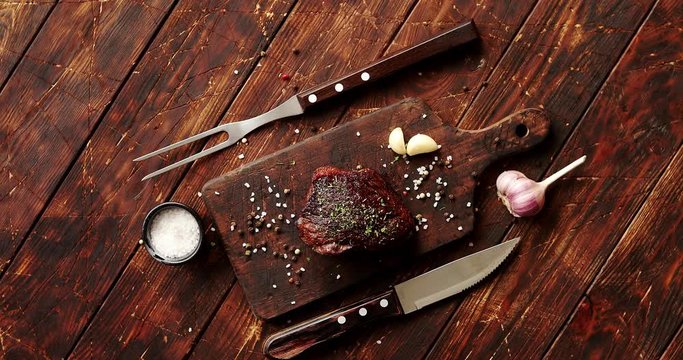 From above view of piece of meat with garlic laid on cutting board with cutlery placed near on wooden background 