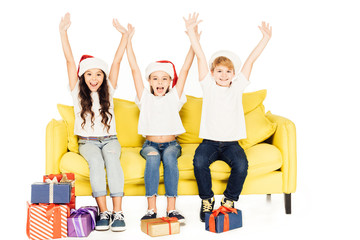 happy adorable kids in santa hats sitting on yellow sofa with raised hands isolated on white