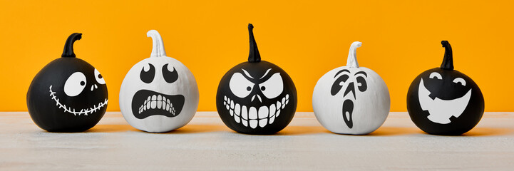 Cute Little Black and White Halloween Pumpkins with funny facial expressions standing side by side on white wooden table over orange background. Halloween banner.