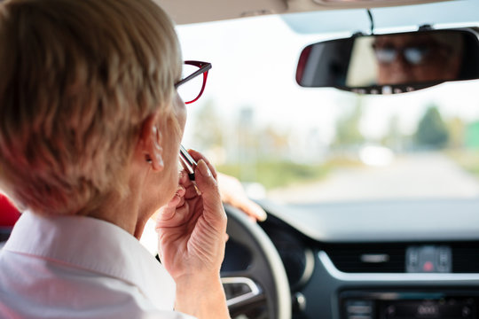 Mature Woman Adjusting Make-up, Putting On Lipstick While Driving A Car. Preparing For Important Meeting. Rear View From Passenger Seat. Commuting By Car Concept.