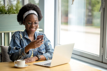 Young african customer client female in spectacles and jeans outfit beaming with joy, spending free time shopping online while sitting in cafe.