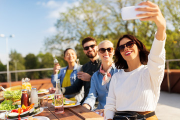leisure and people concept - happy friends with drinks having dinner party on rooftop in summer and taking selfie by smartphone