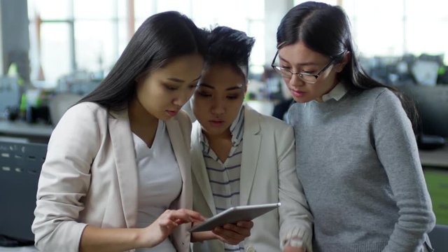 Medium shot of three young Asian women standing close to each other and discussing something they see on tablet computer screen when working together in office