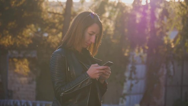 Attractive young girl stands in middle of busy buzzling city on summer sunset. Gorgeous young woman in a bright sunlight uses her phone.