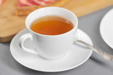 White cup with tea. On a saucer a teaspoon. In the background is an apple pie. Light background. Close-up.