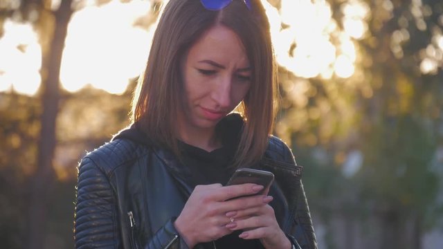Attractive young girl stands in middle of busy buzzling city on summer sunset. Gorgeous young woman in a bright sunlight uses her phone.
