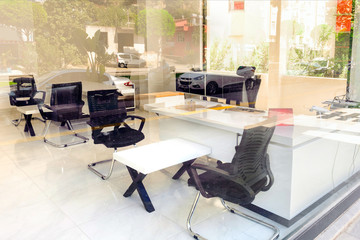 Through the glass wall you can see an empty office with white furniture, the reflection of trees and cars in the glass