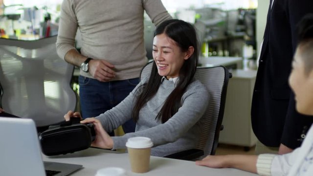 Medium shot of young Asian woman taking off VR headset and giving it to female colleague when having fun with colleagues during break from work in office, handheld