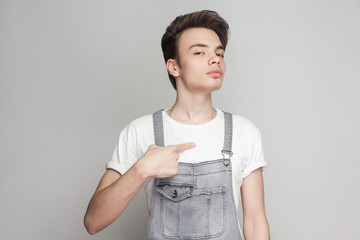 Proud young brunette man in casual style with white t-shirt, denim overalls standing, pointing himself and looking at camera with serious egoist face. indoor studio shot, isolated on gray background.