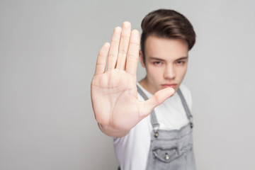 Stop it. Serious young brunette man in casual style with white t-shirt and denim overalls standing and looking at camera with stop or ban gesture. indoor studio shot, isolated on gray background.