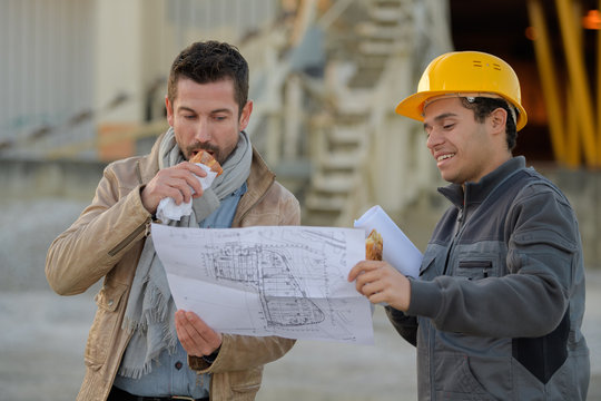 Workmen looking at plans, one eating a sandwich