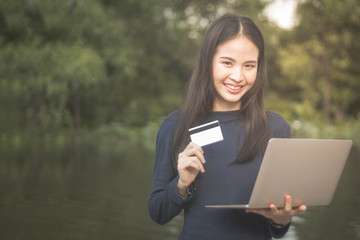 Fototapeta premium Young beautiful Asian woman using credit card and computer laptop for online shoping at outdoor garden park.