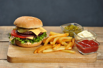 Hamburger on a light wooden surface. The burger includes bacon, fried egg and dried onions. Next to the burger are french fries and sauces. Close-up. 