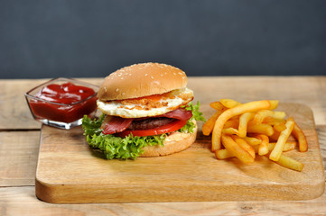 Hamburger on a light wooden board. The burger includes bacon, fried egg and dried onions. Next to the burger are french fries and ketchup cups. Close-up.