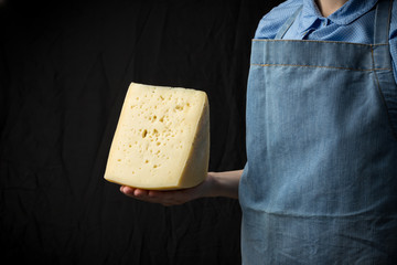 Woman holding slice of cheese wearing blue apron on dark background