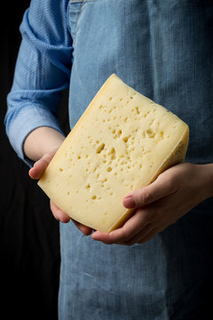 Woman holding slice of cheese wearing blue apron on dark background