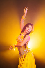 Alluring sexy traditional oriental belly dancer girl dancing on yellow neon smoke background. Woman in exotic costume with feathers sexually moves her semi-nude body