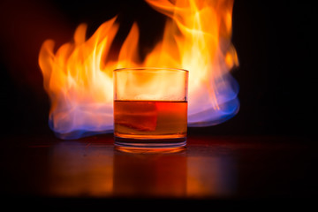 Whiskey in fire concept. Glass of whiskey and ice on wooden surface with color light and fog on background. Close up.
