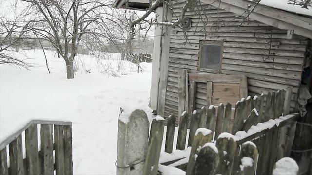 An old dilapidated wooden house with a rickety fence and an orchard near the winter. The concept of loneliness and abandonment