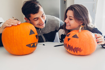 Young couple making jack-o-lantern for halloween on kitchen. Man and woman comparing their pumpkins