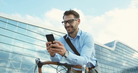 Camera rolling round the smiled caucasian young man in glasses and shirt texting on the smartphone device while sitting on his bike in the city center. Portrait shot. - Powered by Adobe