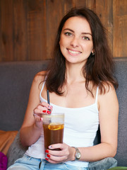 Beautiful cute brunette girl sitting in the cozy cafe near the window enjoying big cup coffee with straw and shy smiling