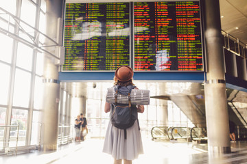 Theme travel public transport. young woman standing with back in dress and hat behind backpack and camping equipment for sleeping, insulating mat looks schedule on scoreboard airport station