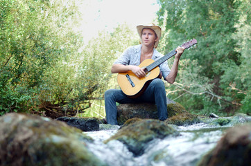 young man playing guitar sitting on a river stone among the turbulent flow of the rapids of the...