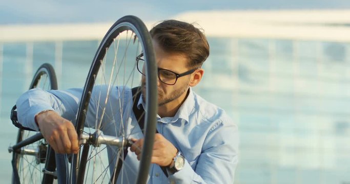 Close up of the handsome Caucasian young man in the casual style scewing the wheel of the bike and checking how it works near the big comercial city center building.