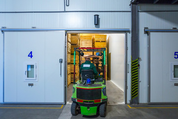 The employee on the electric forklift carry the container wiht ripe apples to inside a fridge airless storage camera. Production facilities of grading, packing and storage of crops of large warehouse. © Sodel Vladyslav